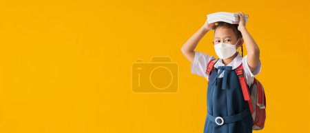 Foto de Back to school banner idea concept, Asian school girl wearing medical face mask with hold books on her head, isolated on yellow background with Clipping paths for design work empty free space - Imagen libre de derechos