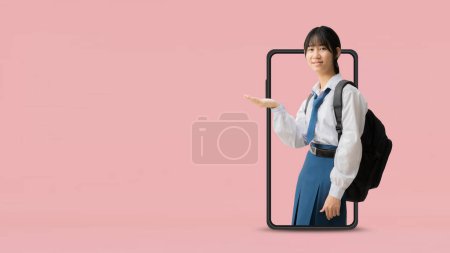 Téléchargez les photos : E learning idea concept, Asian student girl wearing uniform hold backpack with hand holding open palm up coming out big smart phone, isolated on pink background with Clipping paths for design - en image libre de droit
