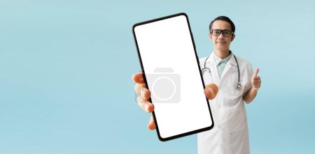 Foto de Asian man doctor coat with hand holding big smart phone, isolated on blue background with Clipping paths for design work empty free space - Imagen libre de derechos