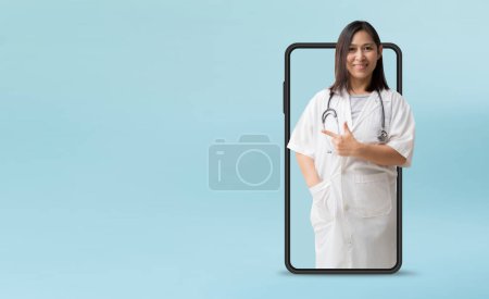 Photo for Medical Services. Smiling asian female doctor pointing finger coming out big smartphone, isolated on blue background with Clipping paths for design work empty free space - Royalty Free Image