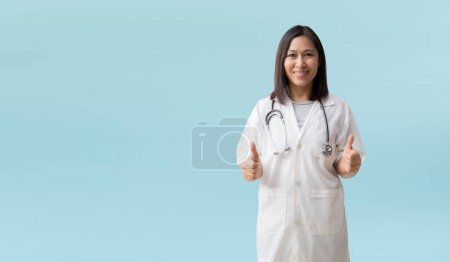 Photo for Positive Caucasian asian female doctor in lab coat showing thumb up gesture, isolated on blue background with Clipping paths for design work empty free space - Royalty Free Image