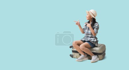 Téléchargez les photos : Pretty asian little girl sits on a suitcase with pointing finger blank space, Adventure vacation travel trip dream concept. isolated on blue background with Clipping paths for design work - en image libre de droit