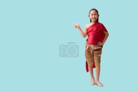 Photo for Full body Asian little girl in Thai national costumes with pointing finger blank workspace, isolated on pastel plain light blue background - Royalty Free Image
