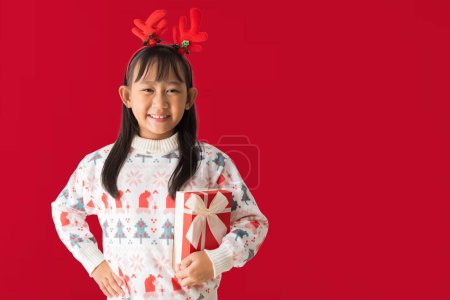 Foto de Alegre niña asiática usando un suéter de Navidad con cuernos de reno, feliz sonrisa mientras sostiene la caja de regalo de Navidad Feliz Año Nuevo, aislado sobre fondo rojo - Imagen libre de derechos