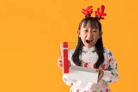 Photo for Cheerful Asian little girl wearing a Christmas sweater with reindeer horns, Happy smiling while holding xmas gift box Happy New Year, isolated on white background, Clipping Paths - Royalty Free Image