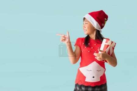 Téléchargez les photos : Joyeux sourire fille asiatique enfant avec le port d'un costume de Noël rouge et le chapeau du Père Noël, Mains pointant la présentation et la tenue boîte cadeau de Noël bonne année, isolé sur fond bleu clair pastel. - en image libre de droit