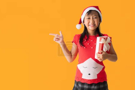Photo for Happy smiling Asian child girl with wearing a red Christmas costume and Santa Claus hat, Hands pointing presentation and holding xmas gift box happy new year, isolated on yellow background - Royalty Free Image