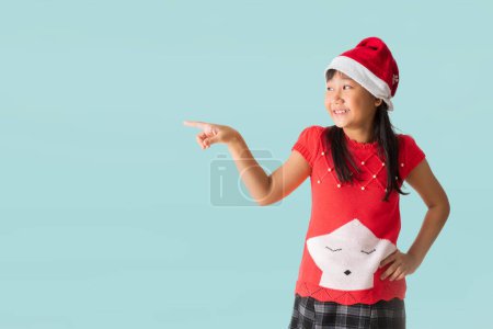 Foto de Feliz niña asiática sonriente con un traje de Navidad rojo y sombrero de Santa Claus, Manos señalando presentación y celebración de la caja de regalo de Navidad feliz año nuevo, aislado en fondo azul claro pastel. - Imagen libre de derechos
