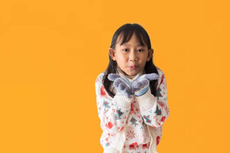 Photo for Portrait of cheerful happy Asian little girl wearing Christmas sweater costume, Hands holding and Sending air kisses posing, isolated on yellow background - Royalty Free Image