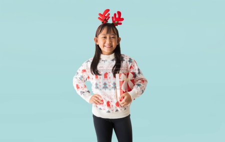 Téléchargez les photos : Portrait de joyeuse petite fille asiatique heureuse portant le costume de pull de Noël, mains tenant boîte cadeau de Noël heureuse nouvelle année, isolé sur fond bleu clair pastel. - en image libre de droit