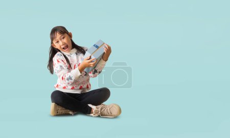 Foto de Retrato de alegre niña asiática feliz con traje de jersey de Navidad, sentado en las manos del piso sosteniendo la caja de regalo de Navidad feliz año nuevo, cuerpo completo aislado sobre fondo azul claro pastel - Imagen libre de derechos