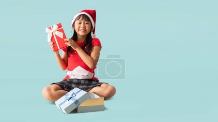 Téléchargez les photos : Portrait de joyeuse petite fille asiatique heureuse femme en santa claus vêtements avec boîte cadeau, mains assises tenant boîte cadeau de Noël bonne année, isolé sur fond bleu clair pastel - en image libre de droit
