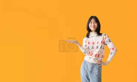 Téléchargez les photos : Positive asiatique fille en chandail de Noël tenir la main démontrer x-mas nouvel an annonce promo toucher doigt porter chaud hiver saison vêtements, isolé sur fond jaune - en image libre de droit