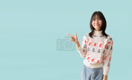 Photo for Happy smiling asian teenager girl pointing at something wearing a Christmas sweater, isolated on pastel plain light blue background - Royalty Free Image