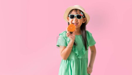 Téléchargez les photos : Joyeux asiatique petite fille posant avec porter un chapeau avec des lunettes de soleil tenant tranches d'orange, vacances été robe verte de mode, isolé sur fond de couleur rose pastel - en image libre de droit
