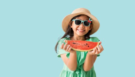 Téléchargez les photos : Joyeux asiatique petite fille posant avec porter un chapeau avec des lunettes de soleil tenant tranches de pastèque, vacances été robe verte de mode, isolé sur fond de couleur bleu pastel - en image libre de droit