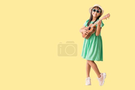 Glücklich lächelnde asiatische Mädchen mit Hut und Sonnenbrille, die Ukulele spielten, versprühten Spaß. Ganzkörper isoliert auf gelbem Hintergrund