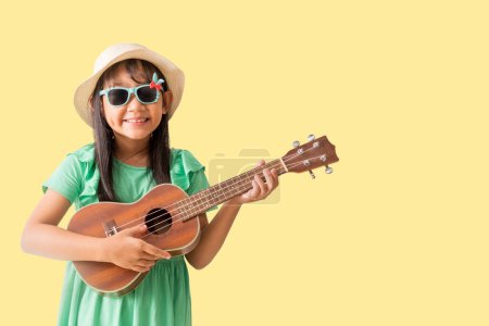 Happy smiling asian little girl were hat and sunglasses playing Ukulele exuding fun. Isolated on yellow background