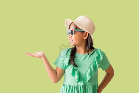 Glücklich lächelnde asiatische Mädchen waren Hut und Sonnenbrille halten Hand Werbung Promo Touch Finger, isoliert auf grünem Hintergrund