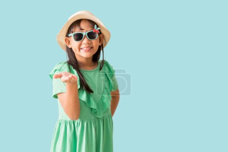 Happy smiling asian little girl were hat and sunglasses hold hand advert promo touch finger, isolated on blue background