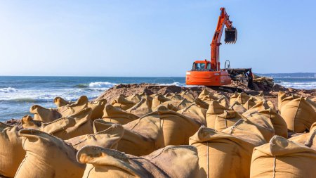 Construcción a plazos con excavadora máquina industrial en la costa de playa de grandes bolsas de arena de lona colocadas como una barrera de protección contra las tormentas de agua de mar y mareas. .
