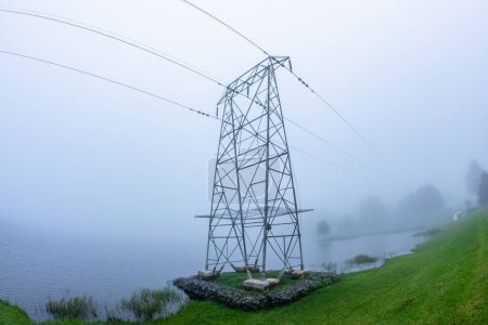 Tour de lignes électriques avec câble haute tension sur le bord de l'eau au-dessus des eaux du barrage dans un paysage nuageux et brumeux.