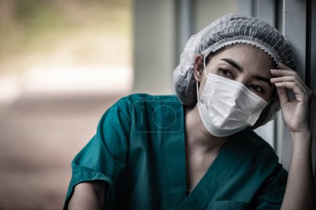 Foto de Cansado deprimido mujer asiática exfoliante enfermera usa mascarilla azul uniforme se sienta en el suelo del hospital, mujer joven médico estresado por el trabajo duro - Imagen libre de derechos