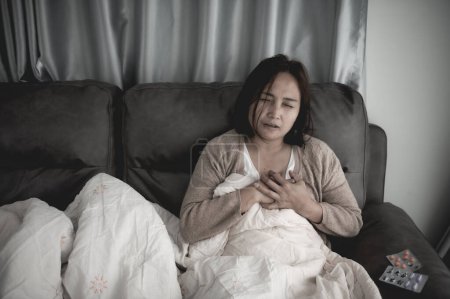 Téléchargez les photos : Asiatique malade femme assise sur le canapé rester à la maison, la femme se sentait mal, voulait se coucher et se reposer, forte fièvre - en image libre de droit