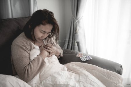 Téléchargez les photos : Asiatique malade femme assise sur le canapé rester à la maison, la femme se sentait mal, voulait se coucher et se reposer, forte fièvre - en image libre de droit