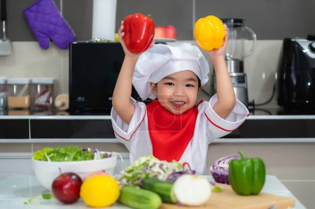 Photo for Cute asian girl wear chef uniform with a lot of vegatable on the table in the kitchen room,Make food for eat dinner,Funny time for kids - Royalty Free Image