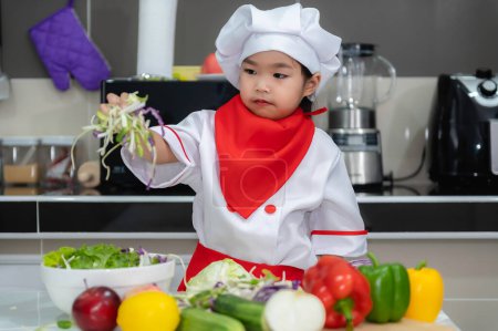 Photo for Cute asian girl wear chef uniform with a lot of vegatable on the table in the kitchen room,Make food for eat dinner,Funny time for kids - Royalty Free Image