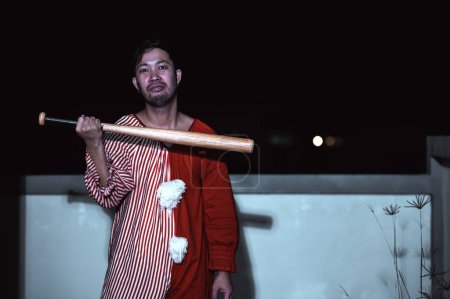 Téléchargez les photos : Asiatique bel homme porter robe de clown avec arme sur la scène de nuit, concept de festival d'Halloween, Horreur photo effrayante d'un tueur en tissu orange, clown maléfique Xotor - en image libre de droit