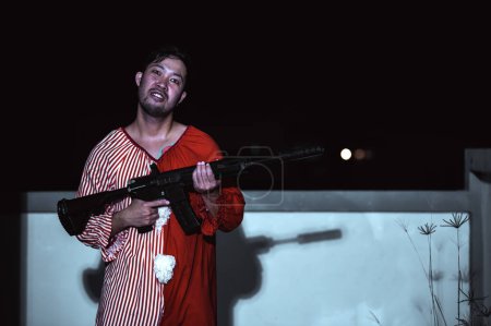 Foto de Hombre guapo asiático usar vestido de payaso con arma en la escena de la noche, concepto de festival de Halloween, Horror aterrador foto de un asesino en tela naranja, Caractor payaso malvado - Imagen libre de derechos