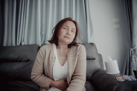 Téléchargez les photos : Asiatique malade femme assise sur le canapé rester à la maison, La femme se sentait mal, voulait se coucher et se reposer, maux d'estomac, douleurs menstruelles - en image libre de droit