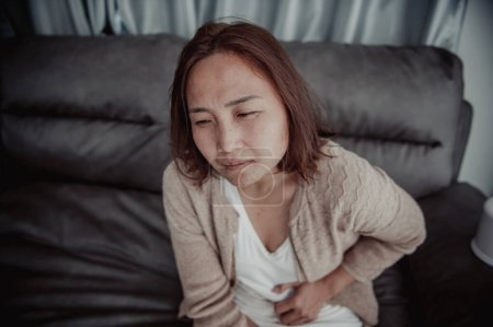 Téléchargez les photos : Asiatique malade femme assise sur le canapé rester à la maison, La femme se sentait mal, voulait se coucher et se reposer, maux d'estomac, douleurs menstruelles - en image libre de droit