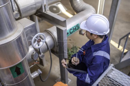Téléchargez les photos : Jeune ingénieur travaillant dans une grande usine, technicien en uniforme de protection et avec contrôle de la température du casque dans les tuyaux - en image libre de droit