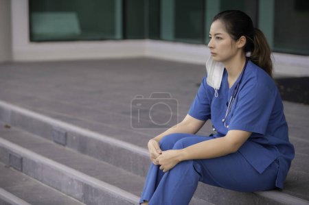 Photo for Tired depressed female asian scrub nurse wears face mask blue uniform sits on hospital floor. Young woman doctor stressed from hard work - Royalty Free Image