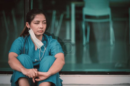 Téléchargez les photos : Fatigué déprimé femme asiatique gommage infirmière porte masque visage uniforme bleu assis sur le sol de l'hôpital. Jeune femme médecin stressé par le travail acharné - en image libre de droit