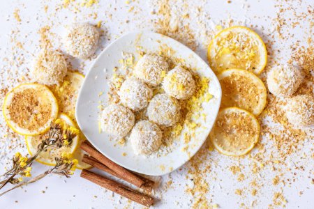 Photo for White chocolate truffles candies with coconut in a white ceramic plate, cinnamon sticks and lemon slices on a white background from above. - Royalty Free Image