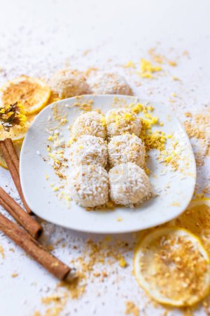 Photo for White chocolate truffles candies with coconut in a white ceramic plate, cinnamon sticks and lemon slices on a white background from above. - Royalty Free Image