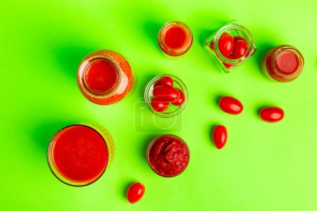 Pequeñas botellas de vidrio de jugo de tomates frescos, tomates cherry, pasta de tomate sobre un fondo verde desde arriba.