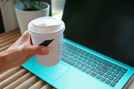 Hand hält eine Tasse mit Getränken und Laptop im Hintergrund
