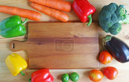 Colorful fresh mixed vegetables surrounding an empty wooden chopping board