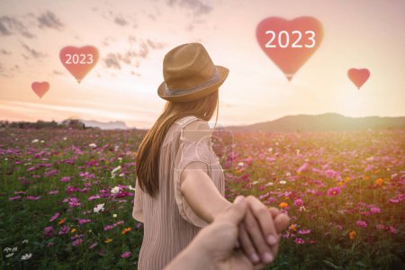 Téléchargez les photos : Jeune voyageur femme tenant la main de l'homme et regardant ballons en forme de coeur volant dans le ciel sur le champ de fleurs, concept de voyage de vacances de couple, concept de célébration du Nouvel An 2023 - en image libre de droit