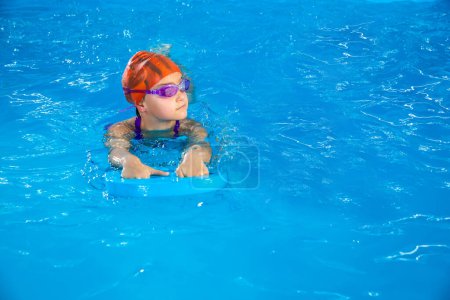 Foto de Little girl learning to swim in indoor pool with pool floating board - Imagen libre de derechos