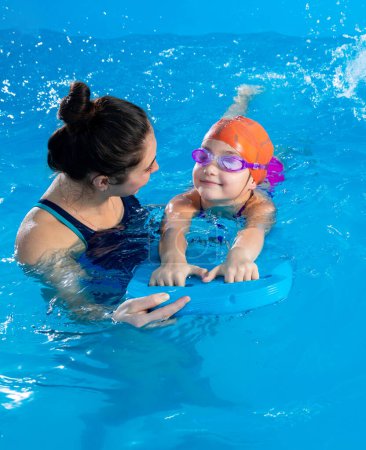 Foto de Little girl learning to swim in indoor pool with pool board. Swimming lesson. Active child swims in water with teacher - Imagen libre de derechos