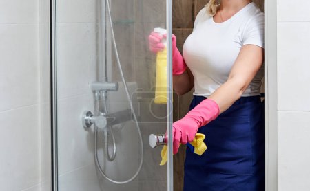 Téléchargez les photos : Jeune belle femme nettoyage cabine de douche en verre avec éponge et vaporisateur - en image libre de droit