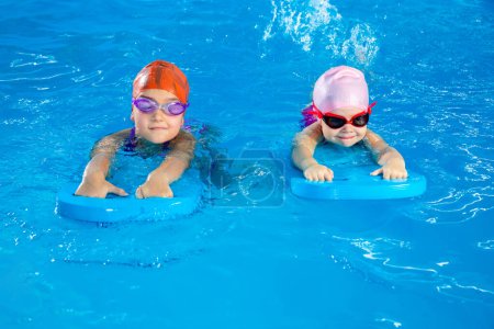Foto de Two little girls learning how to swim in swimming pool and having fun using flutter boards - Imagen libre de derechos