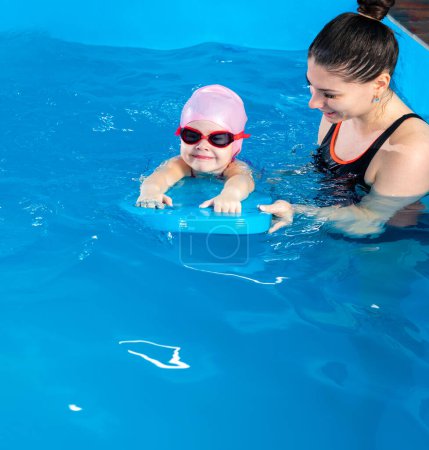 Foto de Little girl learning to swim in indoor pool with pool board. Swimming lesson. Active child swims in water with teacher - Imagen libre de derechos