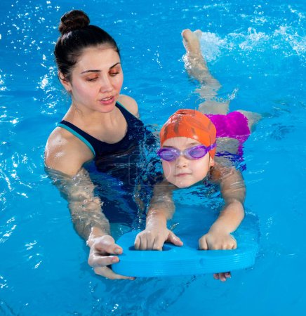 Little girl learning to swim in indoor pool with pool board. Swimming lesson. Active child swims in water with teacher
